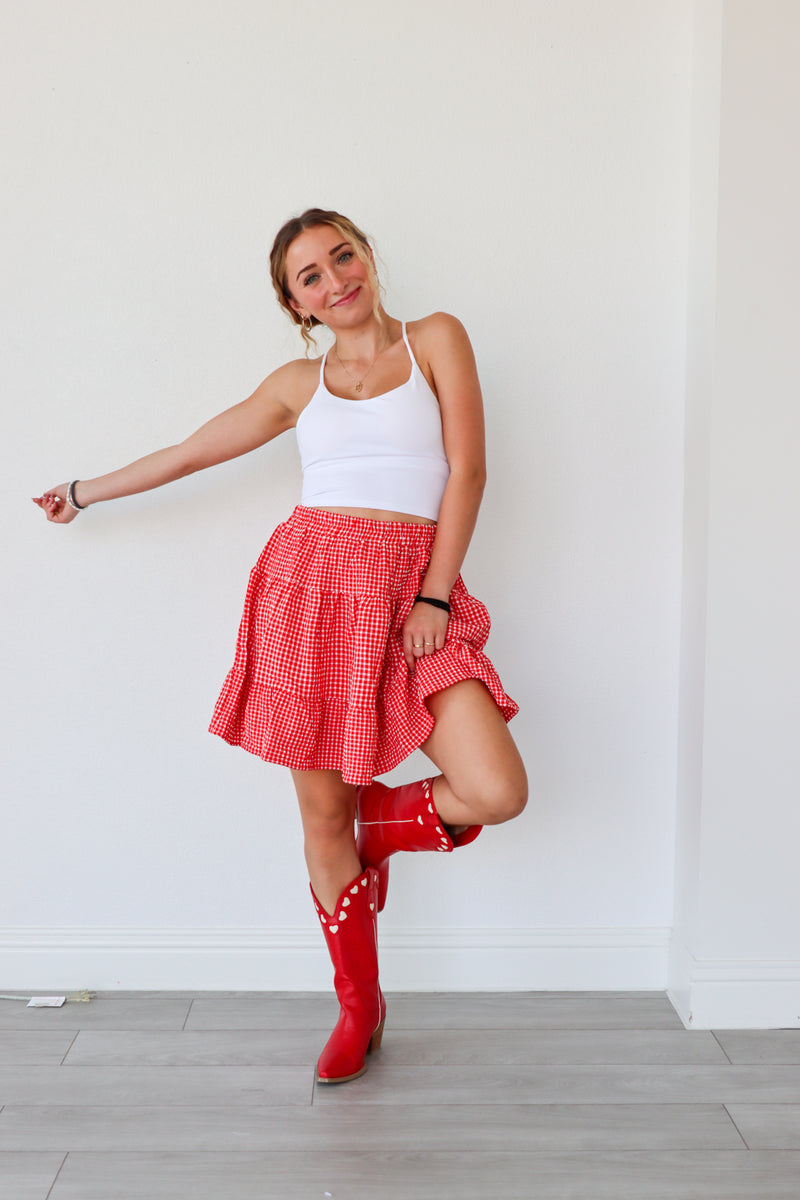 Woman wearing a white top, red checkered skirt, and red rain boots against a white wall.