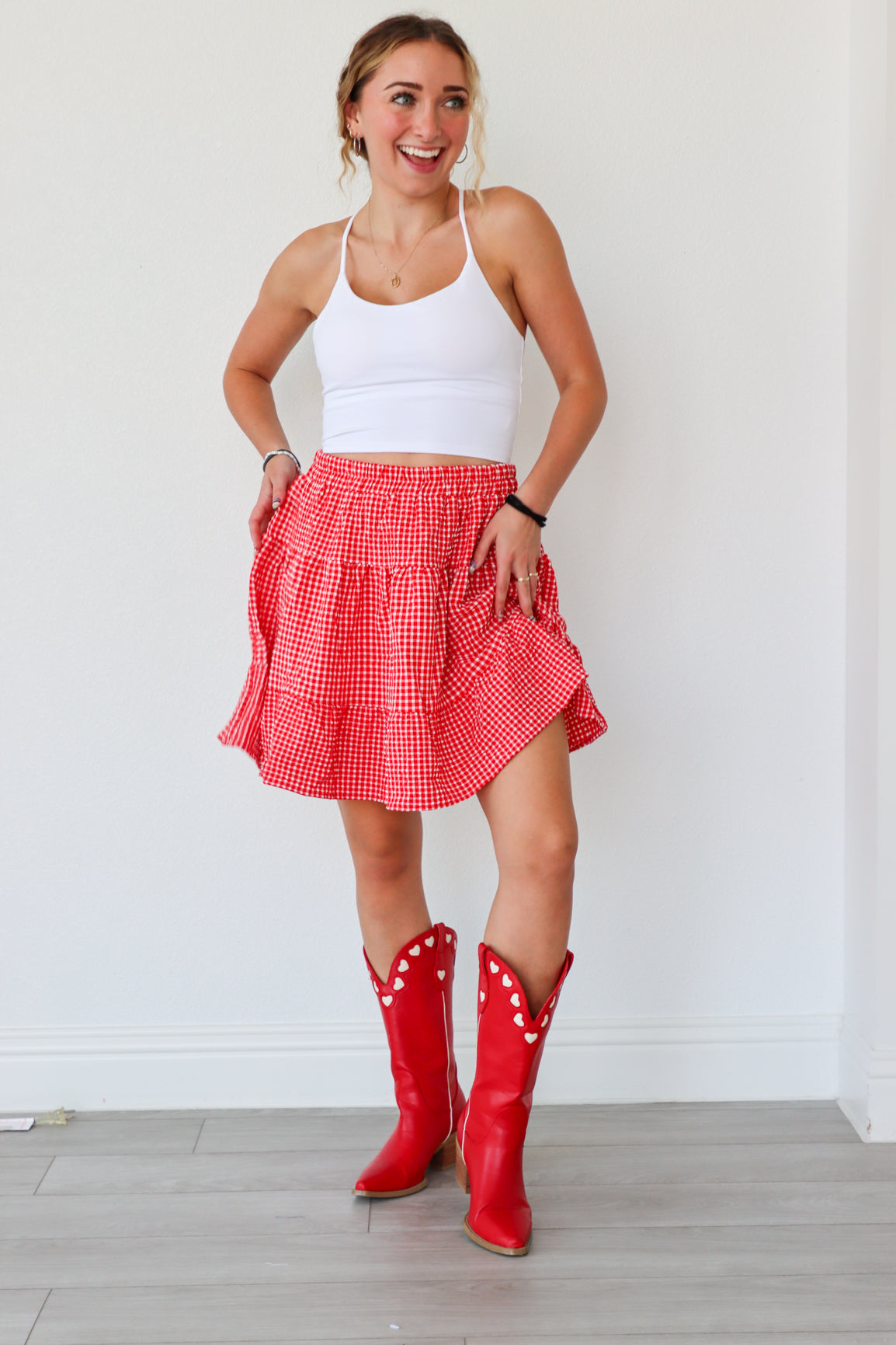 Woman wearing a white tank top, red checkered skirt, and red cowboy boots against a white wall.