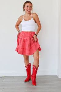 Woman wearing a white tank top, red checkered skirt, and red cowboy boots against a white wall.