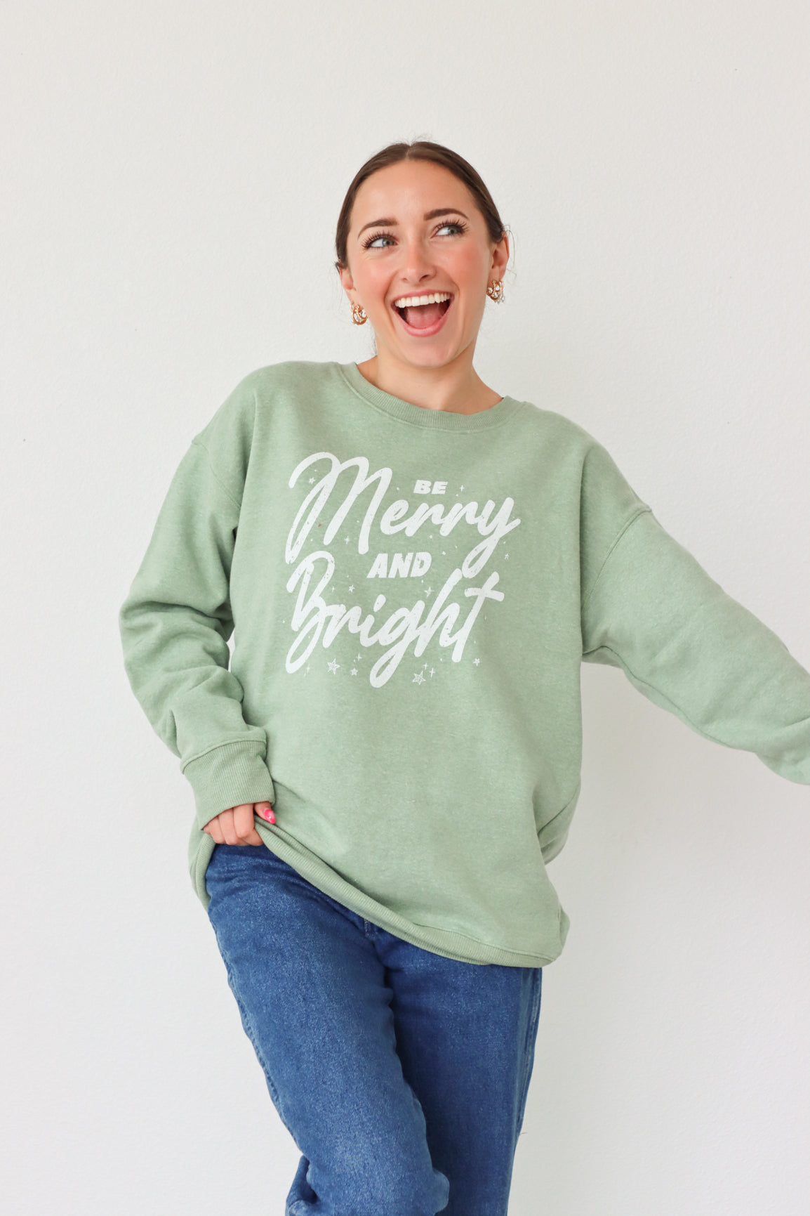 girl wearing light green crewneck sweatshirt with "be merry and bright" graphic