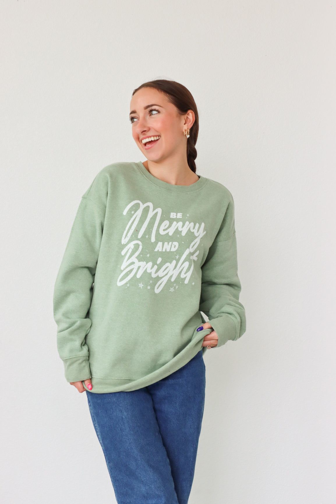 girl wearing light green crewneck sweatshirt with "be merry and bright" graphic