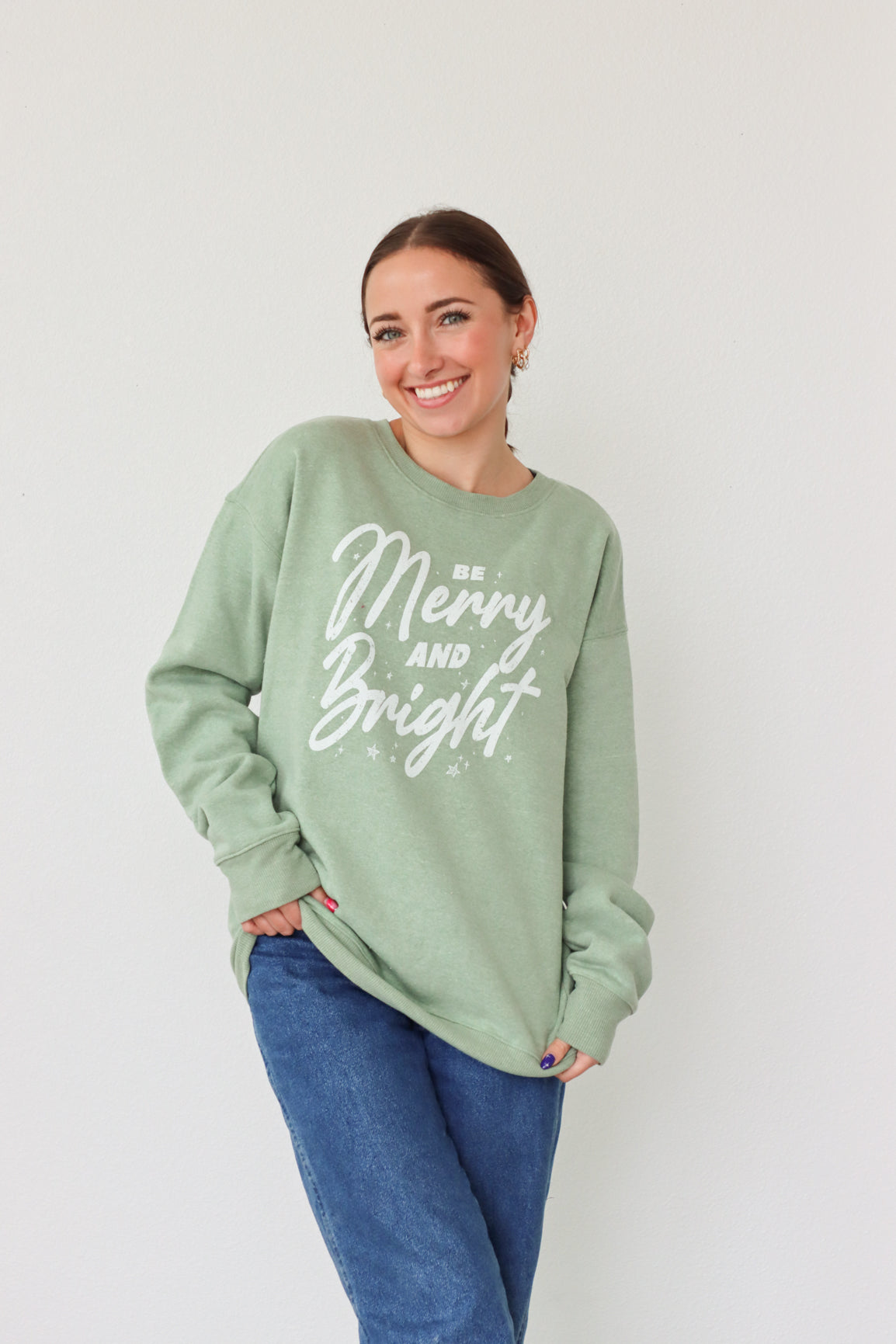 girl wearing light green crewneck sweatshirt with "be merry and bright" graphic