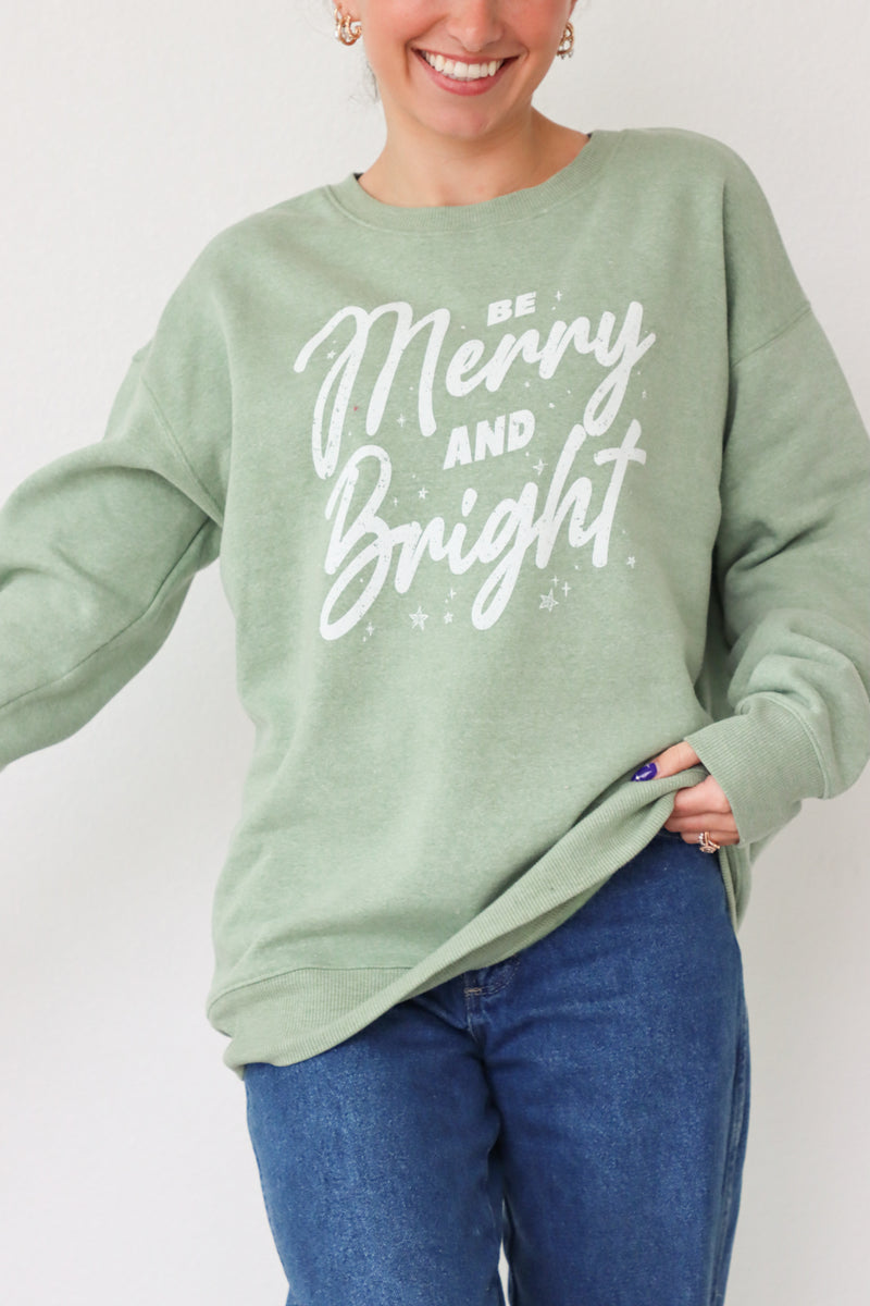 girl wearing light green crewneck sweatshirt with "be merry and bright" graphic