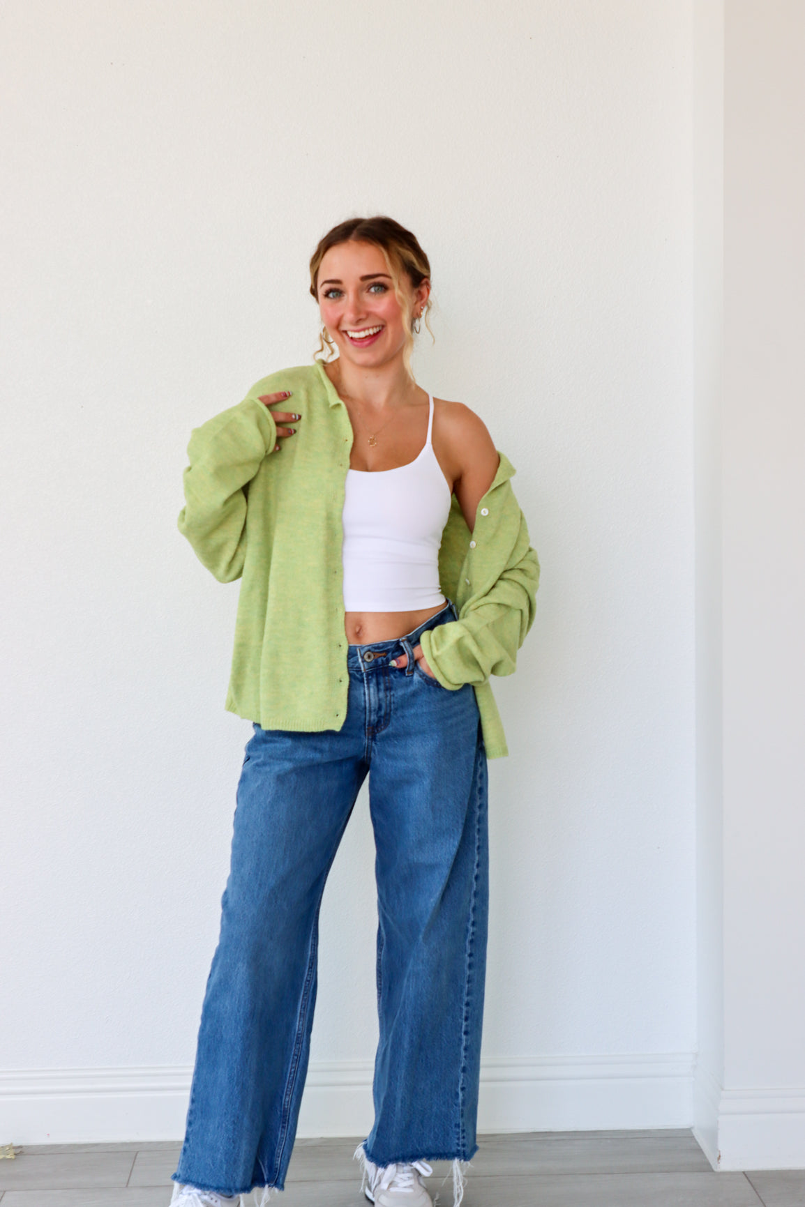 Woman wearing a green cardigan, white top, and blue jeans against a white wall.