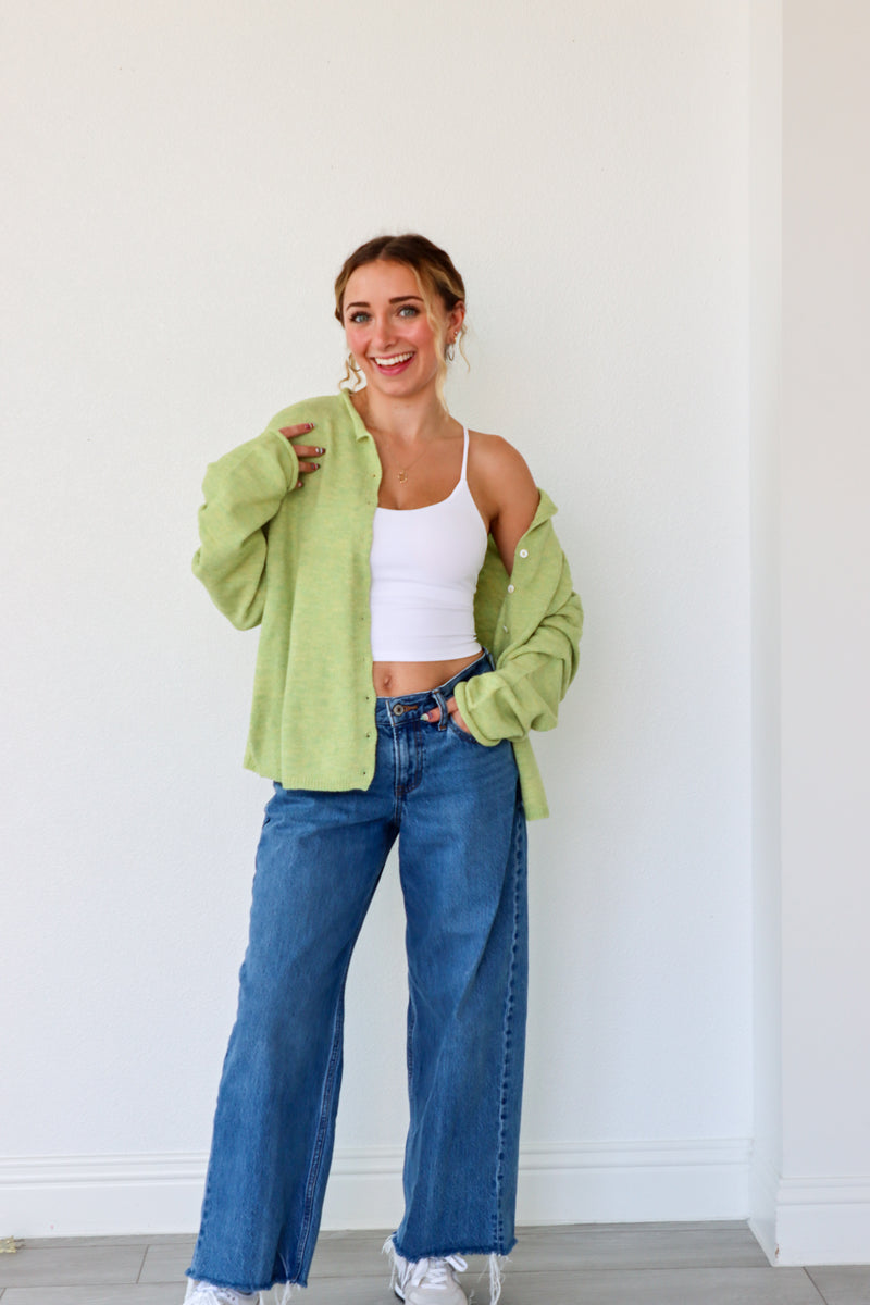 Woman wearing a green cardigan, white top, and blue jeans against a white wall.