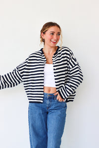 Woman wearing a black and white striped cardigan, white top, and blue jeans against a white background