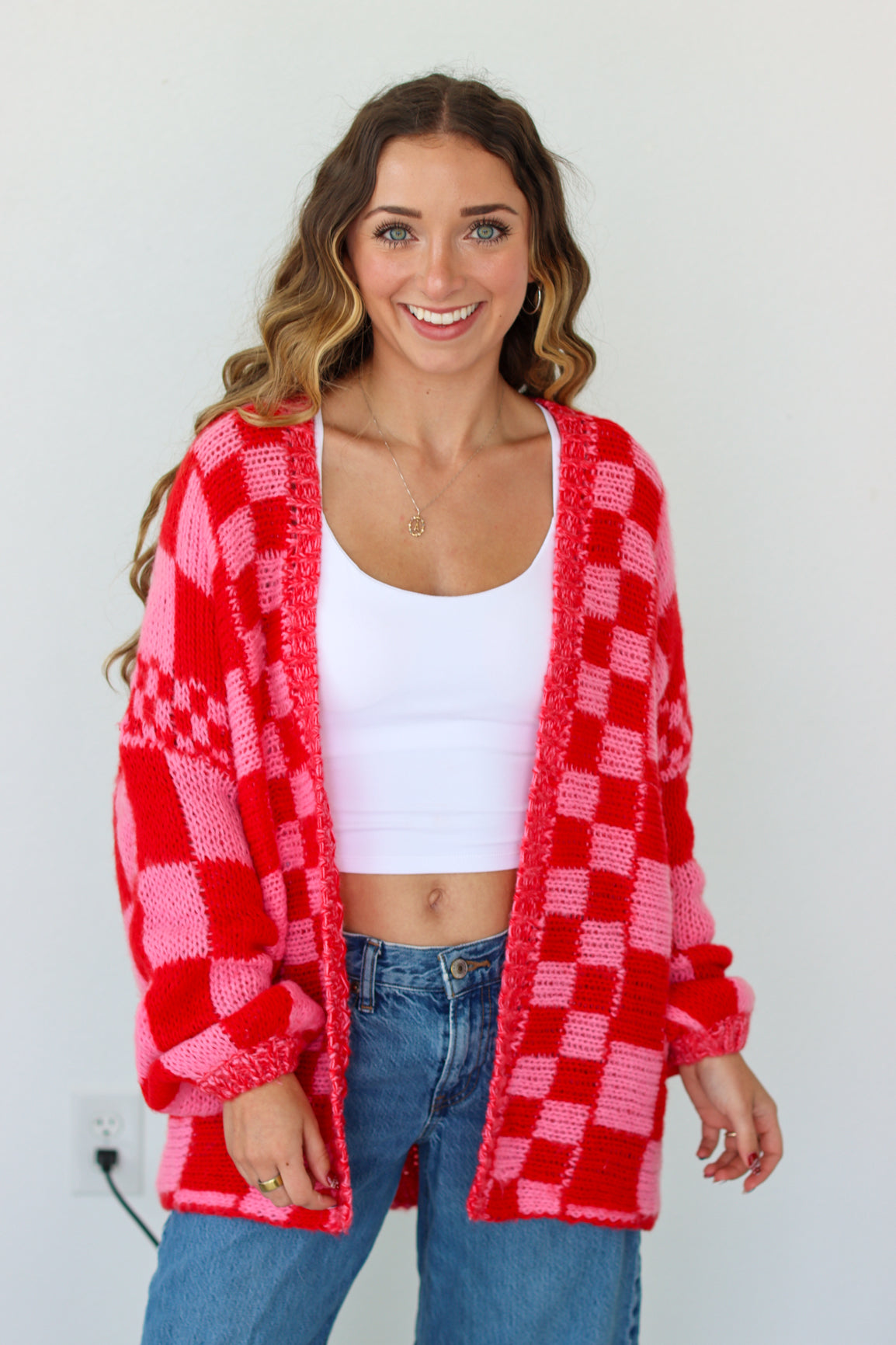Woman wearing a red and pink checkered cardigan over a white top and blue jeans on a plain background