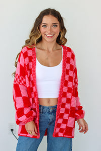Woman wearing a red and pink checkered cardigan over a white top and blue jeans on a plain background