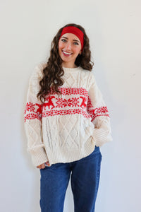 Woman wearing a cream sweater with red deer pattern and blue jeans against a white background