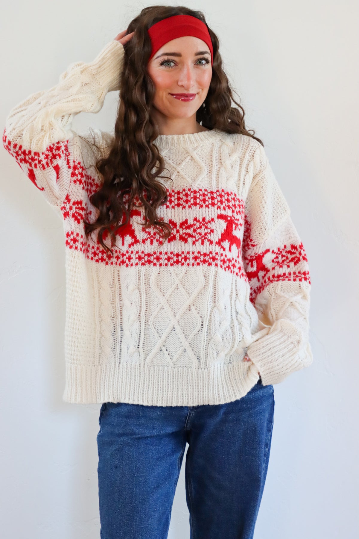 Woman wearing a cream sweater with red and white pattern, blue jeans, and a red headband against a white background.