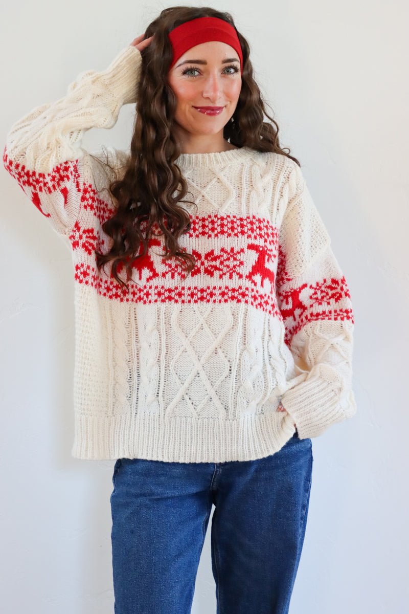 Woman wearing a cream sweater with red and white pattern, blue jeans, and a red headband against a white background.