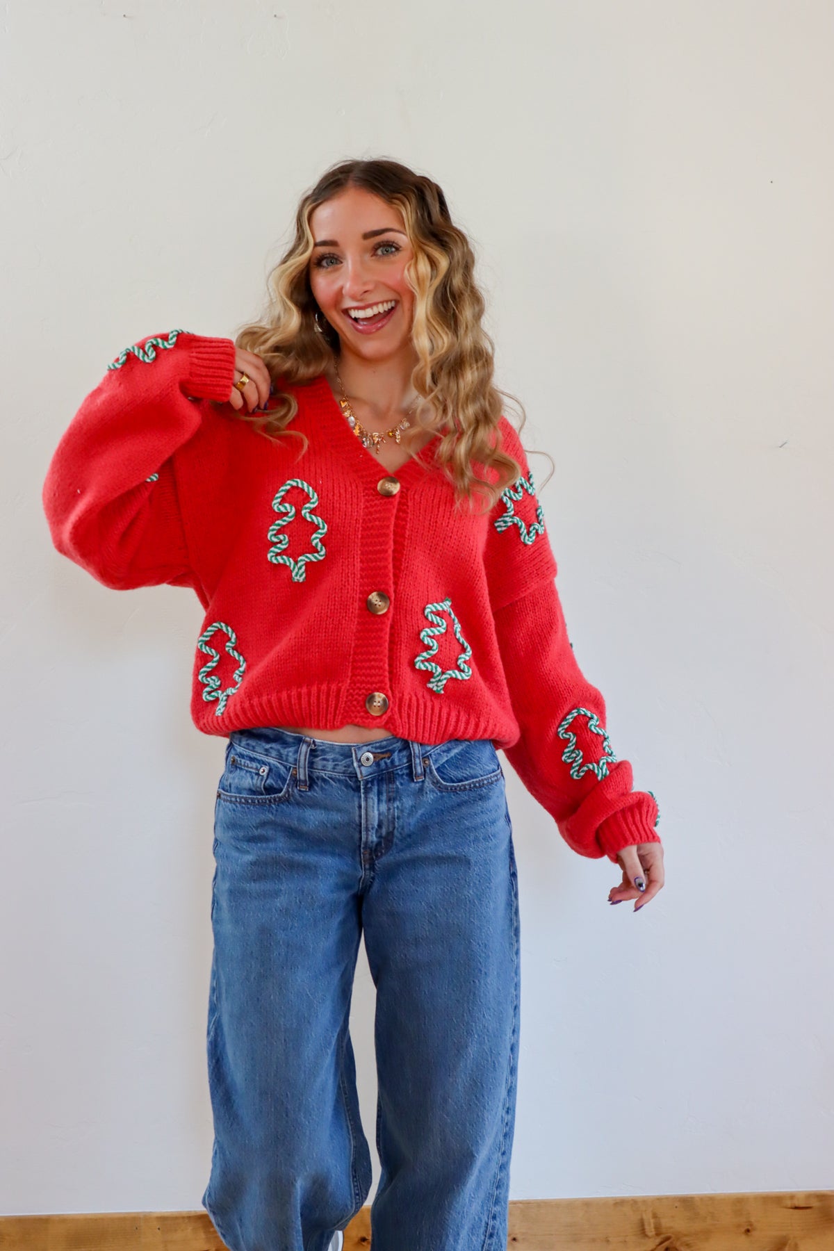 Woman wearing a red sweater with green patterns and blue jeans against a white wall.