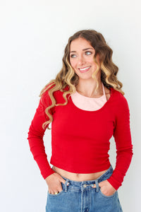 A woman with wavy blonde hair wearing a bright red long-sleeve crop top over a light pink undershirt and blue jeans, standing against a plain white background.