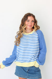 A woman wearing a blue and white striped long-sleeve sweater with solid blue sleeves and a pale yellow ribbed collar, hem, and cuffs, paired with a denim skirt against a plain white background.