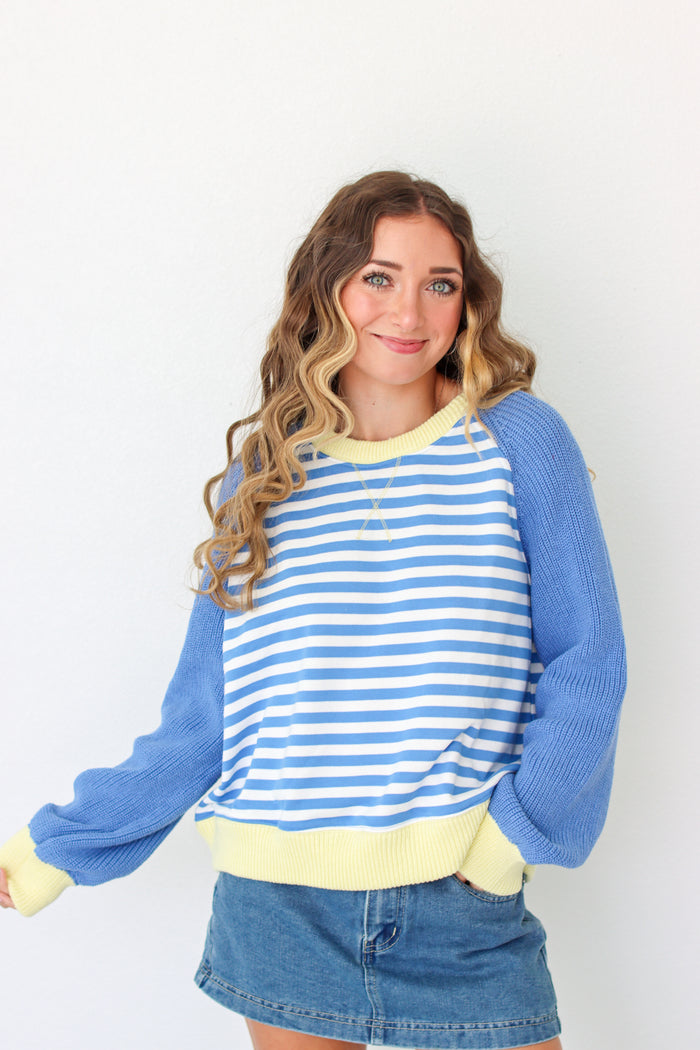 A woman wearing a blue and white striped long-sleeve sweater with solid blue sleeves and a pale yellow ribbed collar, hem, and cuffs, paired with a denim skirt against a plain white background.