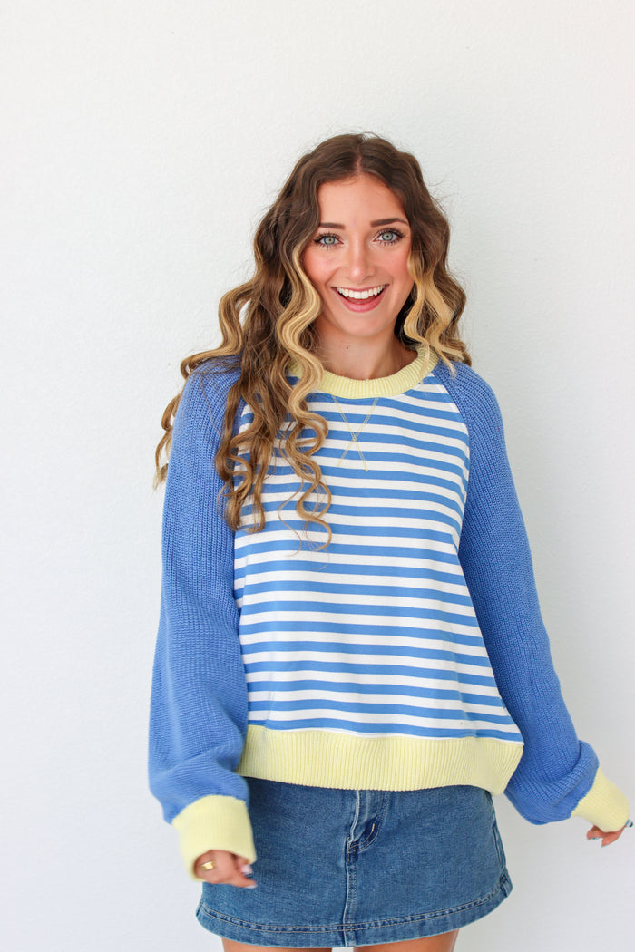 A woman wearing a blue and white striped crewneck sweater with solid blue raglan sleeves and light yellow trim, paired with a denim skirt, against a plain white background.