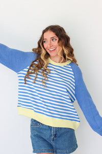 A woman wearing a blue and white striped raglan sweater with yellow ribbed trim, paired with denim shorts, posing against a plain white wall.