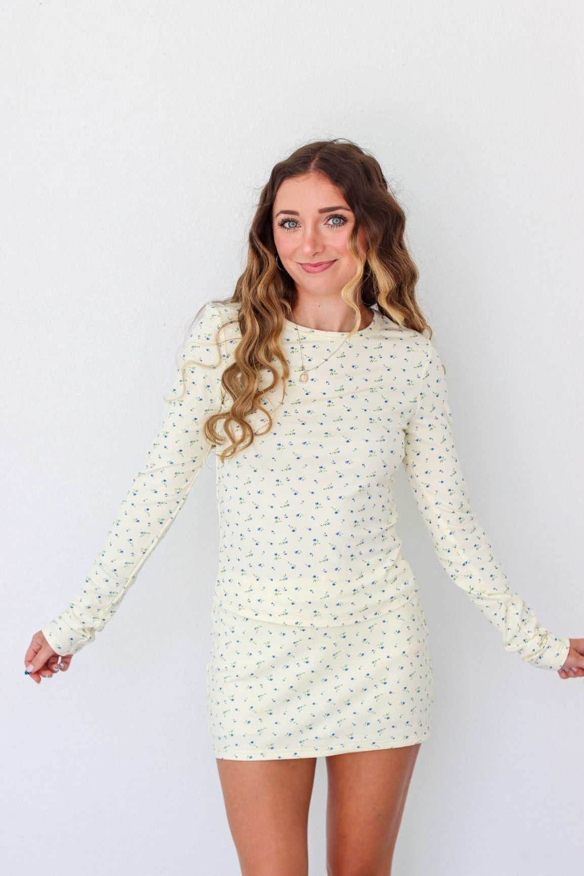 A woman modeling a long-sleeve mini dress with a small, dense all-over print in a light cream or off-white color. The dress has a crew neck and is shown against a plain white studio background.