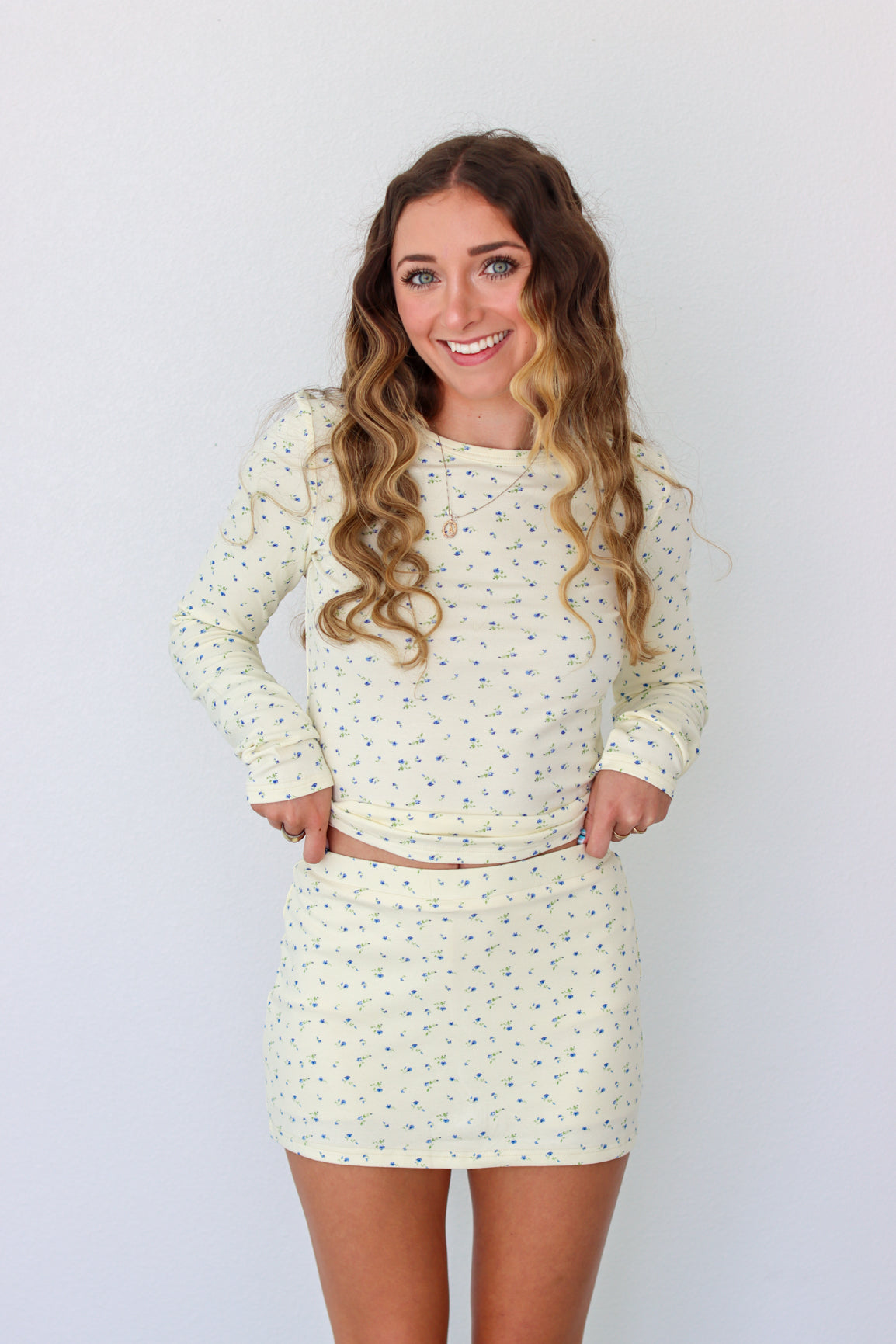 A woman modeling a long-sleeve mini dress with a small, dense all-over print in a light cream or off-white color. The dress has a crew neck and is shown against a plain white studio background.