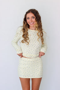 A woman modeling a long-sleeve mini dress with a small, dense all-over print in a light cream or off-white color. The dress has a crew neck and is shown against a plain white studio background.