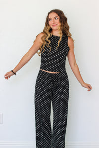 A woman modeling a black sleeveless set with white polka dots, standing against a plain white wall.