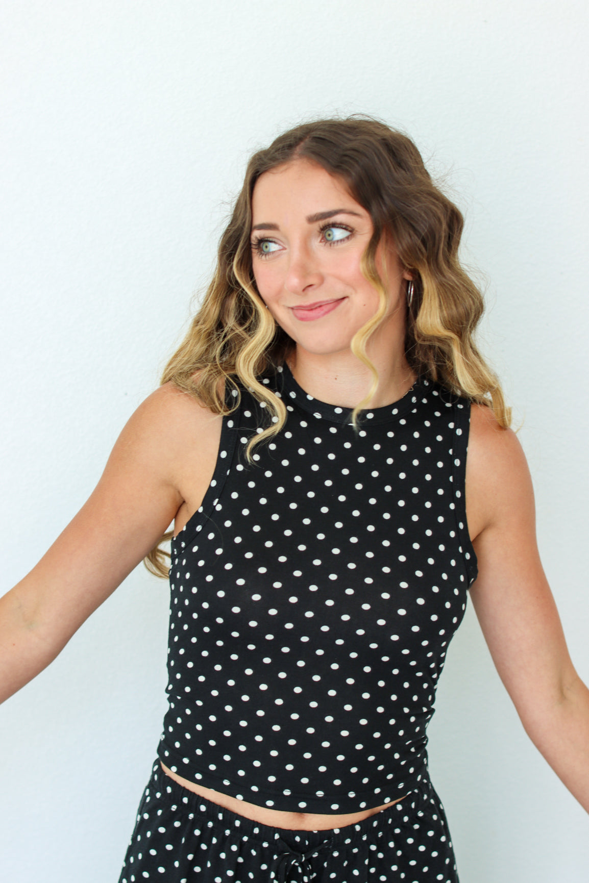A woman modeling a black sleeveless set with white polka dots, standing against a plain white wall.