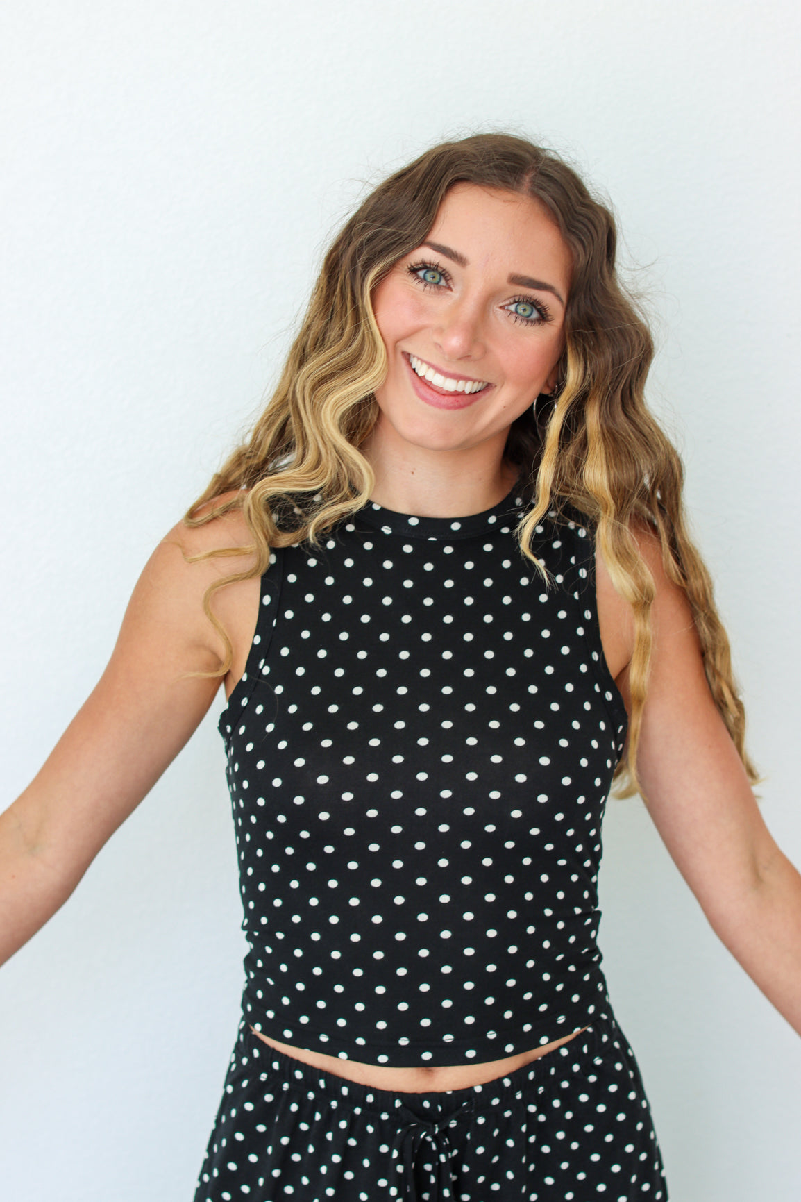 A smiling woman with long wavy brown and blonde hair wearing a black sleeveless crop top and matching black polka dot pants, posed against a plain light gray background.