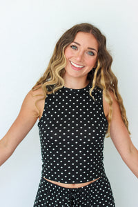 A smiling woman with long wavy brown and blonde hair wearing a black sleeveless crop top and matching black polka dot pants, posed against a plain light gray background.