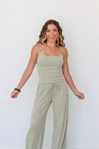 A woman modeling a light sage green and yellow striped camisole set with a drawstring waist, standing against a plain white wall.