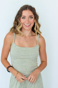 A smiling woman with wavy blonde-highlighted hair wearing a light sage green and yellow striped spaghetti-strap tank dress, standing against a plain light blue background.