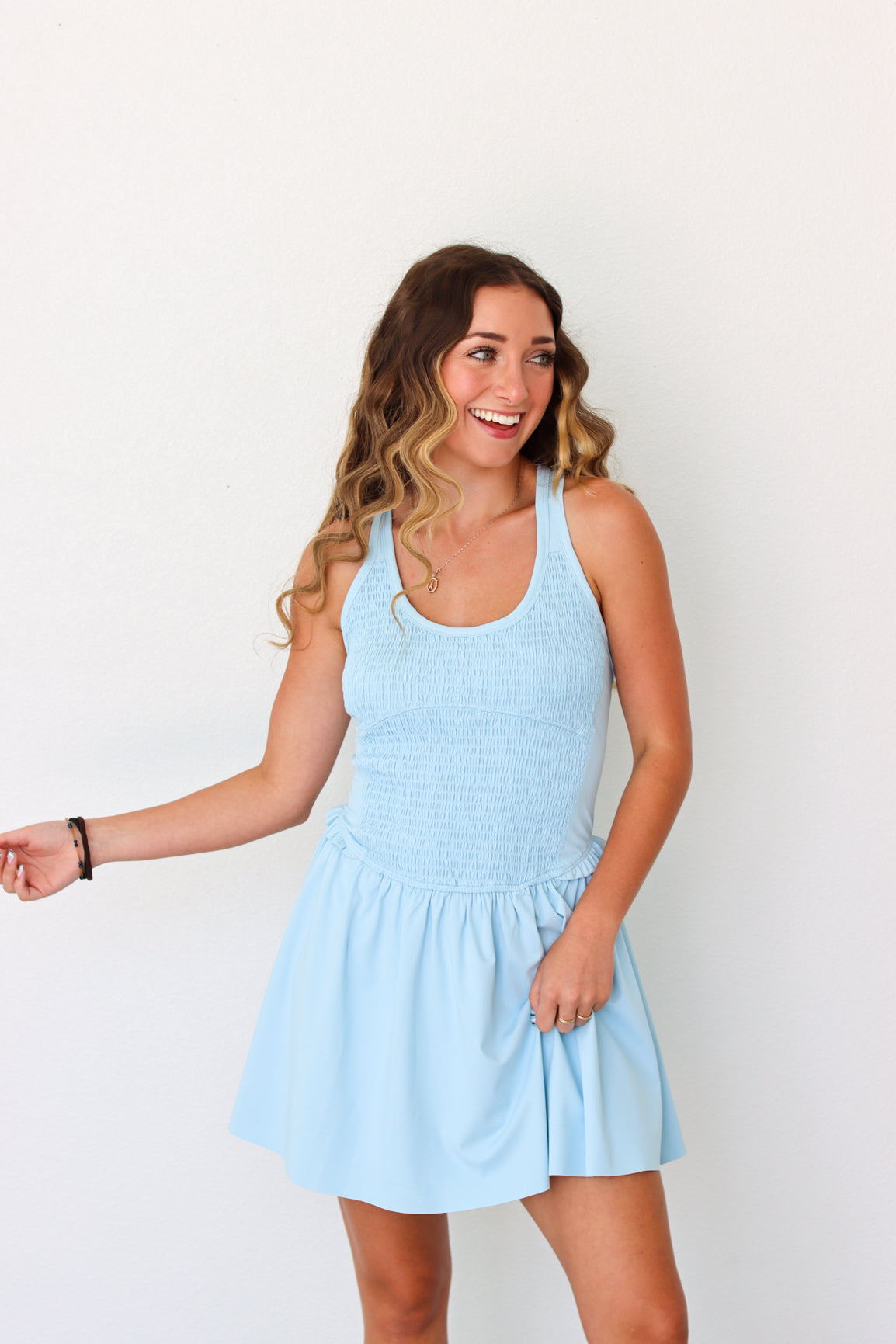 A woman modeling a light blue sleeveless mini dress with a smocked bodice and flared skirt, standing against a plain white background.