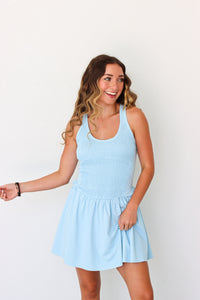 A woman modeling a light blue sleeveless mini dress with a smocked bodice and flared skirt, standing against a plain white background.