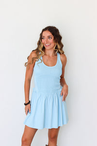 A woman modeling a light blue sleeveless mini dress with a smocked bodice and flared skirt, standing against a plain white background.