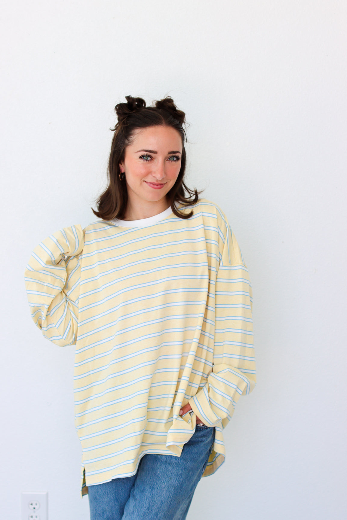 A woman with her hair in two buns wears a pale yellow and white striped long-sleeve top with blue jeans, standing against a plain white wall.