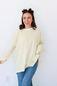 A woman with her hair in two buns is wearing a yellow and white striped long-sleeve t-shirt and blue jeans, standing against a plain white wall.