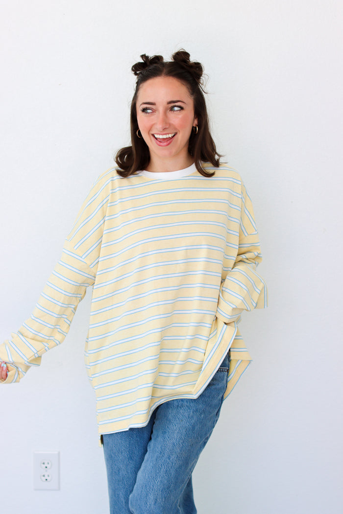 A woman with her hair in two buns is wearing a yellow and white striped long-sleeve t-shirt and blue jeans, standing against a plain white wall.