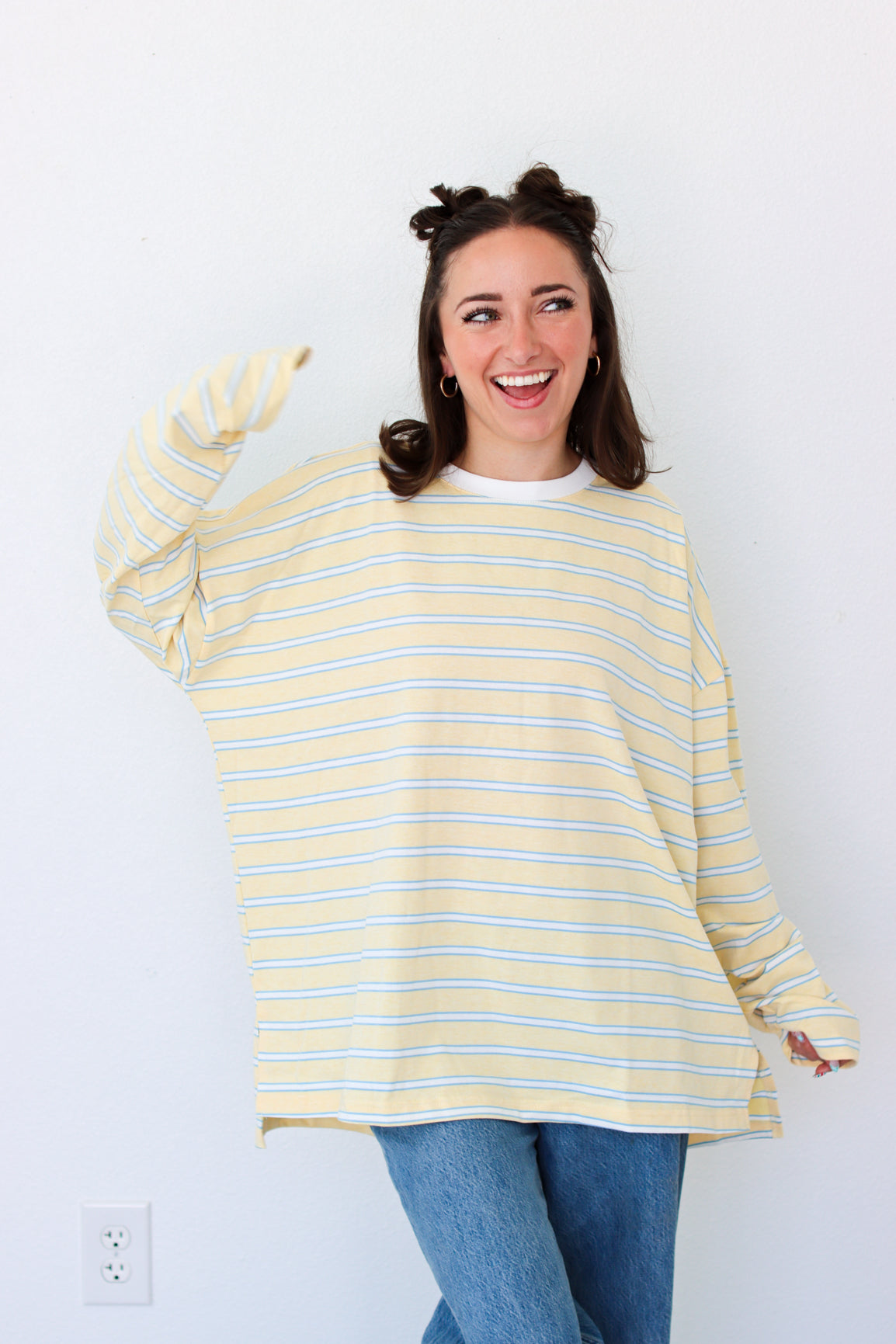 A woman wearing a yellow and white striped long-sleeve t-shirt and blue jeans, smiling and pointing towards the camera against a plain white wall.