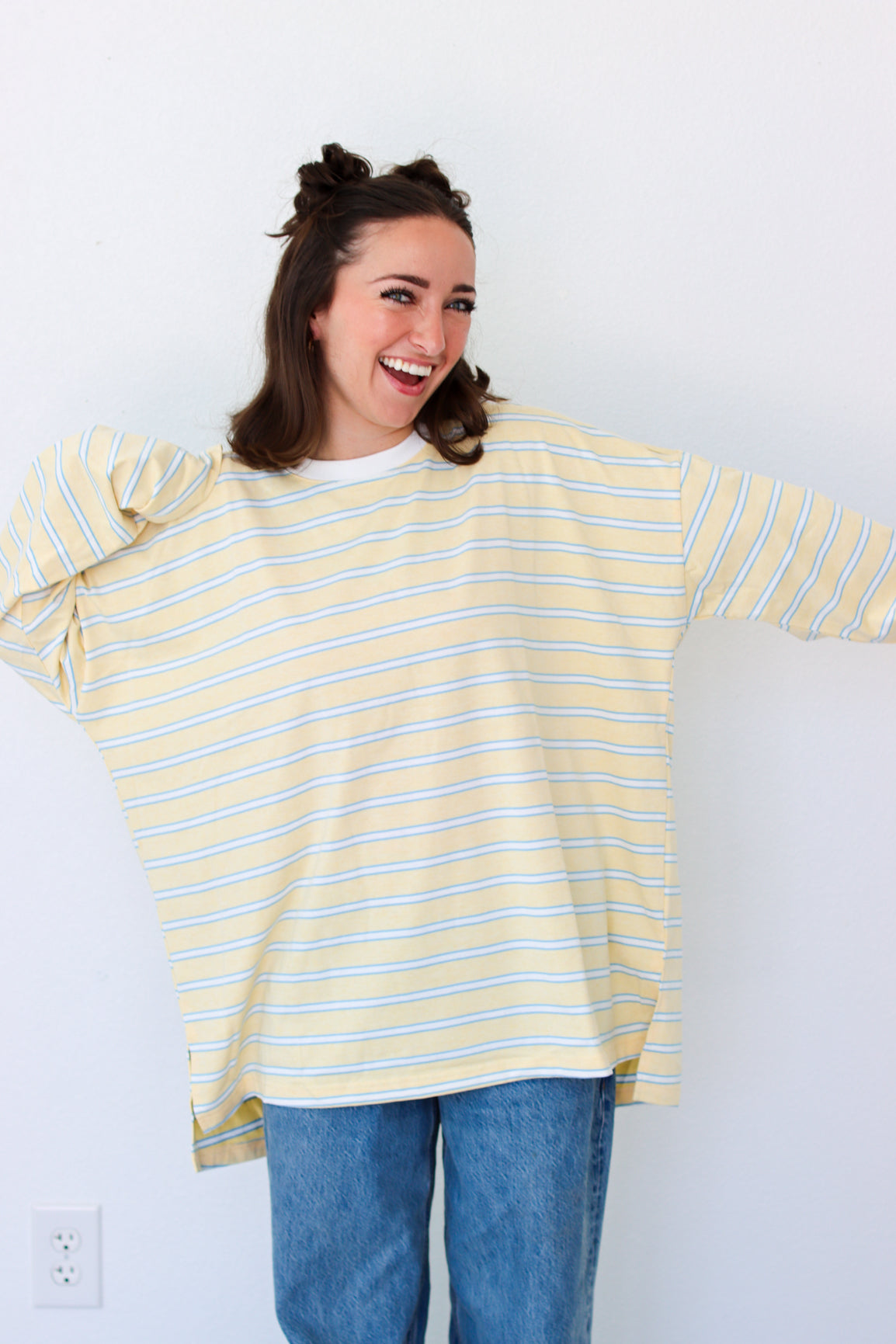 A woman wearing a yellow and white striped oversized long-sleeve shirt with blue jeans, posing against a plain white wall.
