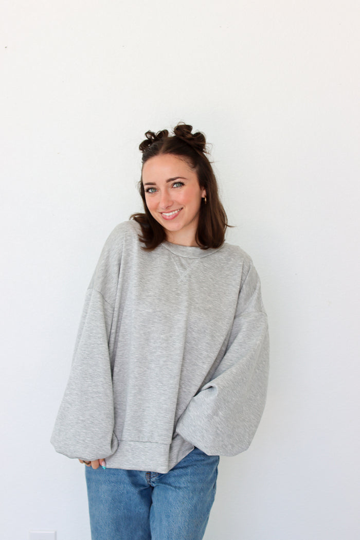 A woman with her hair in two buns wears an oversized light gray heather crewneck sweatshirt and blue jeans, standing against a plain white wall.