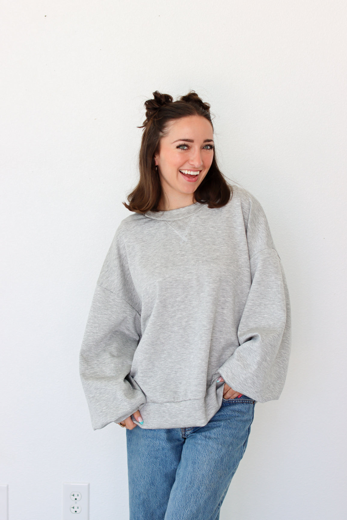 A woman smiling and wearing an oversized light gray crewneck sweatshirt with blue jeans, standing against a plain white wall.
