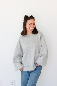 A woman smiling and wearing an oversized light gray crewneck sweatshirt with blue jeans, standing against a plain white wall.