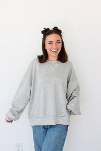 A woman smiling and wearing an oversized light gray heather crewneck sweatshirt with blue jeans, standing against a plain white wall.