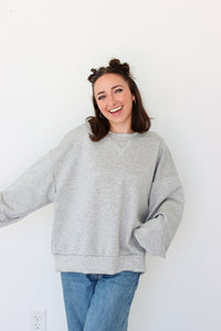A woman smiling and wearing an oversized light gray heather crewneck sweatshirt with blue jeans, standing against a plain white wall.