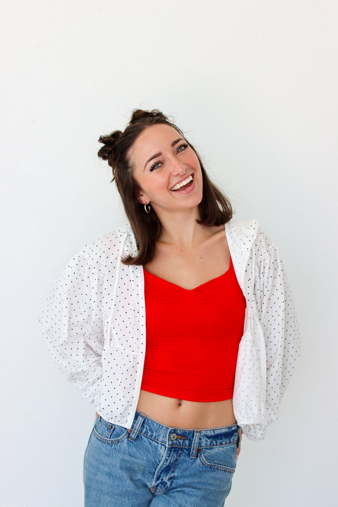 A woman wearing a bright red crop top, a white open-front sheer polka dot jacket, and blue jeans, smiling against a plain white background.