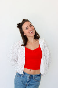 A woman wearing a bright red crop top, a white open-front sheer polka dot jacket, and blue jeans, smiling against a plain white background.