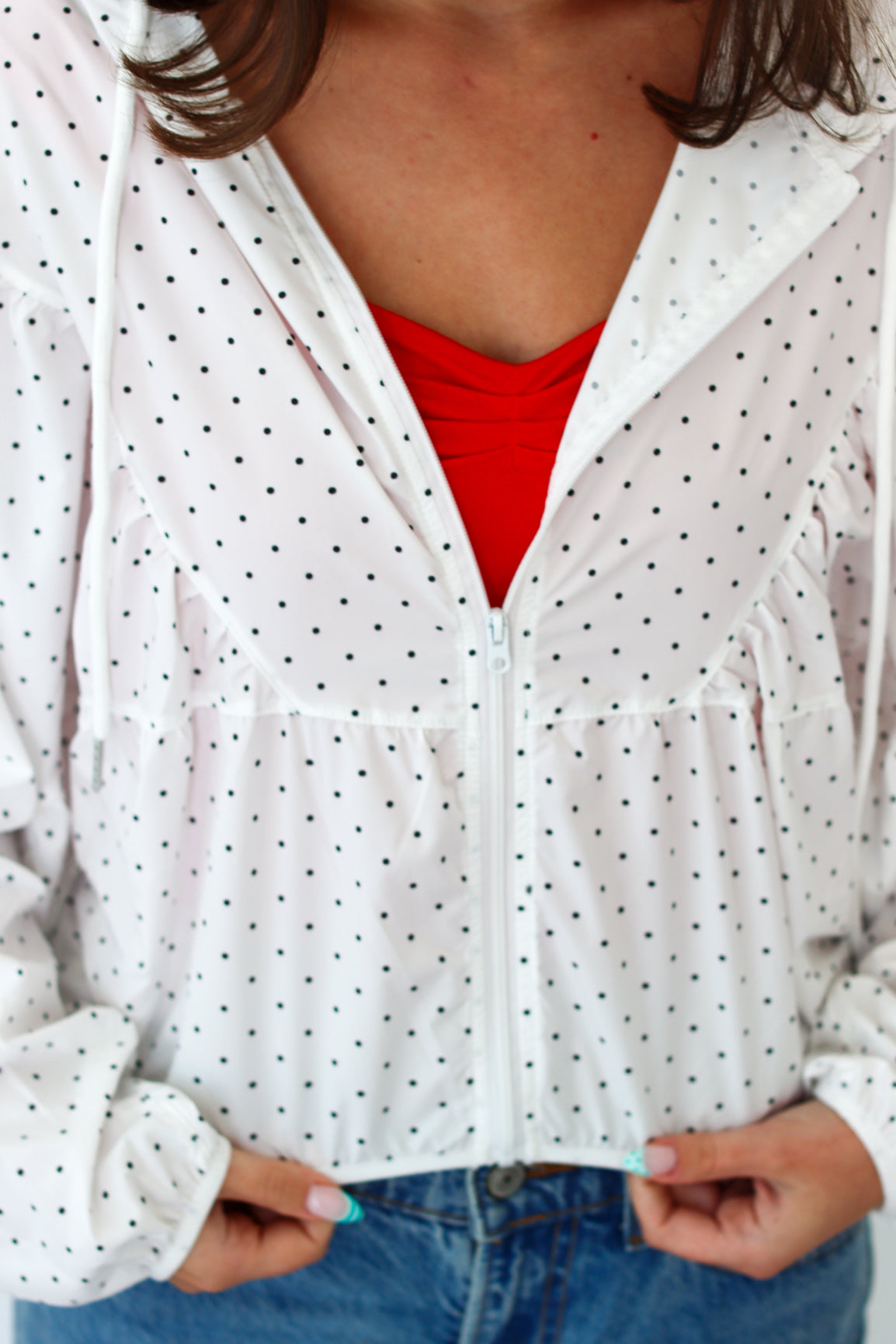A close-up of a person wearing a white zip-up hoodie with small black polka dots, layered over a bright red top and paired with blue jeans.