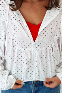 A close-up of a person wearing a white zip-up hoodie with small black polka dots, layered over a bright red top and paired with blue jeans.