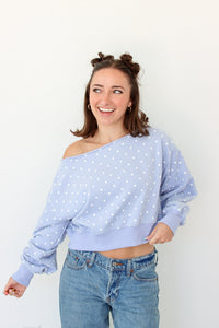 A woman wearing a light blue off-the-shoulder long-sleeve sweatshirt with white polka dots and blue jeans, standing against a plain white wall.