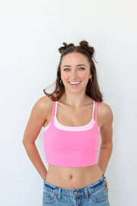 A smiling woman with her hair in two buns wears a bright pink crop top with a light pink trim, paired with blue jeans, standing against a plain white wall.