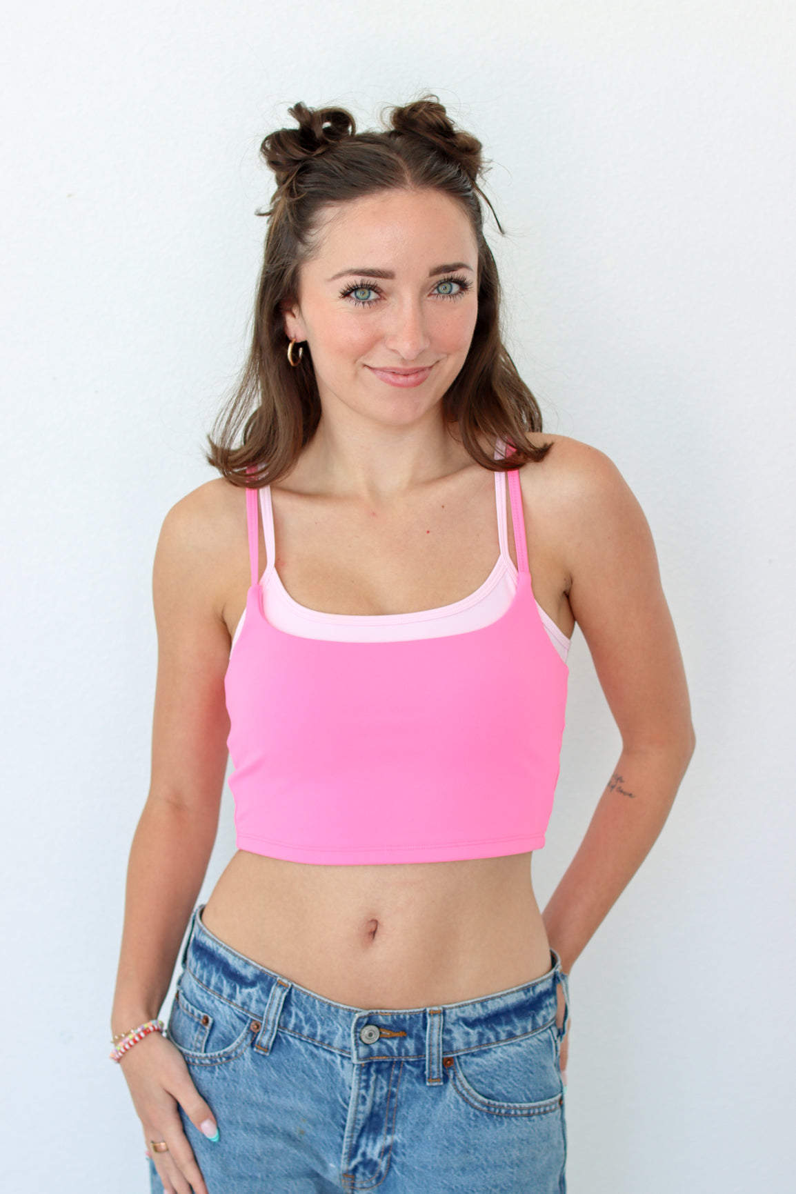 A woman modeling a bright pink crop top with a contrasting light pink scoop neckline, paired with blue jeans, against a plain white background.